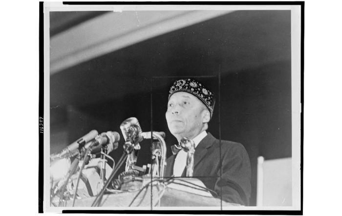 Elijah Muhammad stands behind microphones at a podium in 1964.